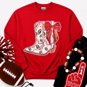 May include: A bright red crewneck sweatshirt featuring a large central graphic of a white cowboy boot adorned with sequined footballs, gold stars, and a prominent red sequined bow. The sweatshirt has a Gildan brand label at the inner neckline.