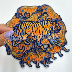 May include: Embroidered tiger patches in orange, yellow, and blue. The patches feature a detailed tiger design with a navy blue border. A hand holds one patch, with several more patches in the background.