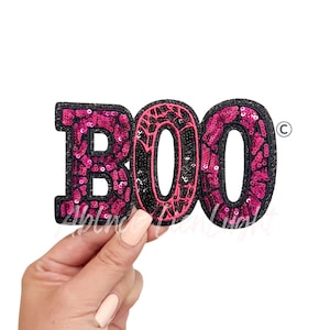 May include: A sequined patch spelling "BOO" in pink and black. The letters are outlined in black glitter and filled with pink sequins. The letter "O" in the middle has a spiderweb design. The patch is held by a hand.