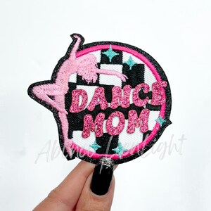 May include: Pink and black iron-on patch with a silhouette of a dancer and the text "Dance Mom" in pink glitter lettering. The background is a black and white checkered pattern with turquoise stars.