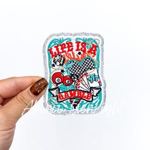 May include: Embroidered patch with a silver glitter border. The patch features a turquoise background with the words "Life Is A Gamble" in red. Images of dice, playing cards, and billiard balls are included.