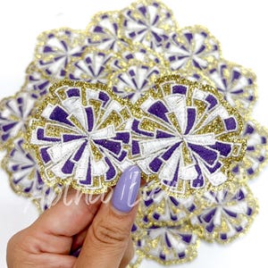 May include: A close-up of a pile of purple, white, and gold glitter iron-on patches in the shape of a cheerleading pom-pom.
