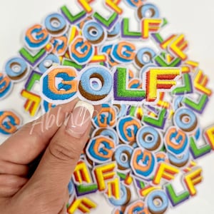 May include: A pile of colorful embroidered iron-on patches spelling out the word "GOLF". The patches are in various colors, including blue, orange, yellow, green, and purple.