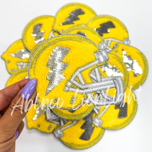 May include: A pile of yellow and silver iron-on patches featuring a football helmet with a lightning bolt design. The patches are made of sequins and are perfect for adding a sporty touch to clothing or accessories.