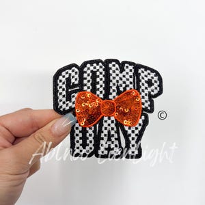 May include: A black and white checkered fabric patch with the word "GAME DAY" in black lettering and an orange sequined bow tie.
