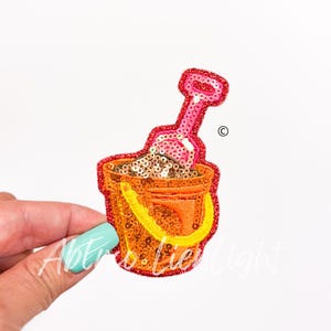 May include: A colorful, sequined iron-on patch of a bucket and shovel. The bucket is orange with yellow trim and filled with brown sand. The shovel is pink with a white handle.