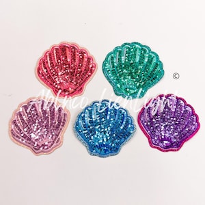May include: Five different colored fabric seashells with a sparkly finish. The shells are pink, teal, purple, blue, and light pink.