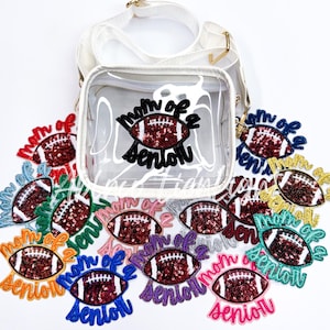May include: A clear plastic crossbody bag with a white strap. The bag features a patch that reads "Mom of a Senior" with a red and gold football inside. There are several other patches with the same design in different colours around the bag.