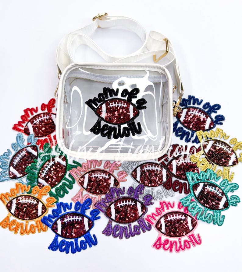 May include: A clear plastic crossbody bag with a white strap. The bag features a patch that reads "Mom of a Senior" with a red and gold football inside. There are several other patches with the same design in different colours around the bag.