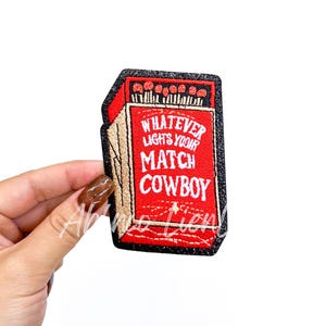 May include: Embroidered patch in the shape of a matchbox, featuring the text "WHATEVER LIGHTS YOUR MATCH COWBOY" in white on a red background. The patch has a black glitter border and a tan side.