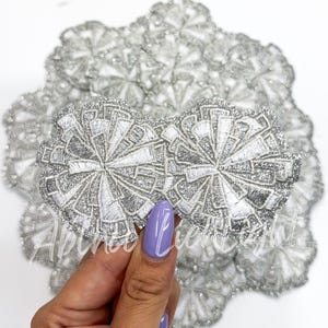 May include: A close-up of a silver and white embroidered flower applique. The applique is made of a sparkly fabric and has a delicate, intricate design.