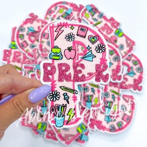 May include: Pink felt iron-on patch with the word "PRE-K" in glitter lettering. The patch is decorated with school-themed illustrations, including a backpack, apple, pencil, and a cup of pencils.