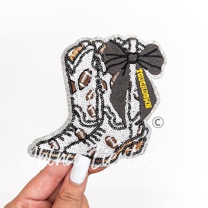 May include: A sequined patch shaped like a cowboy boot featuring a white background with brown football patterns and a large black bow. The word TOUCHDOWN is embroidered in yellow on the side of the boot. The patch has a silver glitter border.