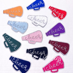May include: Twelve iron-on patches in various colors featuring a megaphone with the word "cheer" written on it. The patches are decorated with silver glitter.