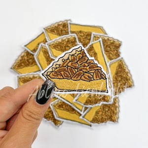 May include: A set of 10 iron-on patches featuring a slice of pecan pie. The patches are a light brown color with a dark brown filling and a light brown crust. The patches are perfect for adding a touch of whimsy to your clothing, bags, or other accessories.