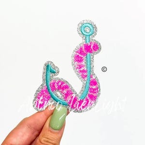 May include: A decorative, sequined fish hook-shaped patch. The patch features a bright pink sequined body with a teal blue outline and accents. The edges are outlined with silver glitter. The patch is on a white background.