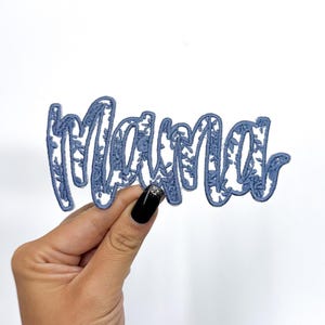 May include: A blue decorative cutout of the word "Mama" with floral detailing. The script is held against a white background. The cutout is a light blue color.