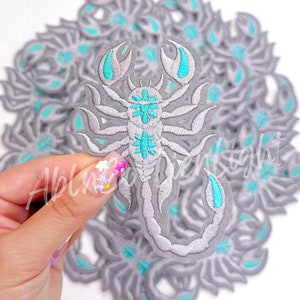 May include: A pile of gray embroidered iron-on patches featuring a scorpion design with turquoise accents.