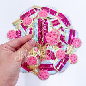 May include: A pile of pink and blue sequin patches with a gold outline. The patches are shaped like paddles and balls, and are perfect for adding a touch of fun to any project.