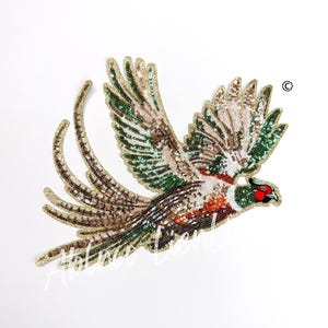 May include: A green, brown, and gold sequined pheasant applique. The pheasant is in flight with its wings spread and tail feathers trailing behind it. The pheasant has a red head and a black eye.