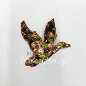 May include: A gold, green, and brown camouflage patterned sequined iron-on patch of a flying duck.
