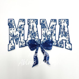 May include: A white and blue floral design spelling out the word "MAMA" with a navy blue sequined bow. The letters are outlined in navy blue and filled with a white fabric featuring a blue floral pattern. The bow is made of navy blue sequins.