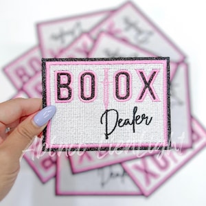 May include: A stack of white and pink iron-on patches with black embroidery. The top patch features the text "BOTOX Dealer" in black and pink lettering.