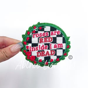 May include: A round, green, glittery patch with a black and white checkered background. The patch has red roses around the edge and the text "roses are red inside i am dead" in red.