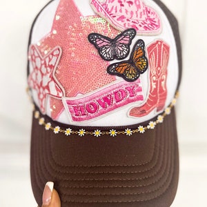 Puede incluir: Gorra de camionero marr&oacute;n y blanca con visera marr&oacute;n y panel frontal blanco. La parte delantera presenta una estrella de lentejuelas rosa, sombrero de vaquero, bota de vaquero y parches de mariposa. La palabra "HOWDY" est&aacute; bordada en una pancarta rosa.