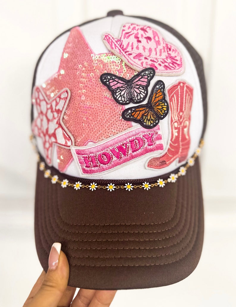 Puede incluir: Gorra de camionero marr&oacute;n y blanca con visera marr&oacute;n y panel frontal blanco. La parte delantera presenta una estrella de lentejuelas rosa, sombrero de vaquero, bota de vaquero y parches de mariposa. La palabra "HOWDY" est&aacute; bordada en una pancarta rosa.