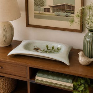 May include: A rectangular ceramic tray with a pale, glossy, light green glaze and curved, flared edges. The tray sits on a wooden surface and contains a small metal key ring and a single green leafy sprig.