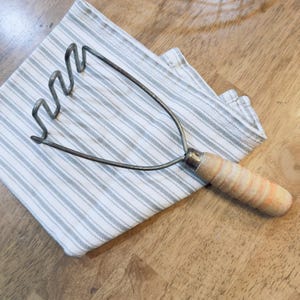 May include: A vintage potato masher with a wooden handle and metal mashing head rests on a striped gray and white kitchen towel. The handle is light-colored wood with orange stripes.