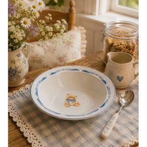 Op de afbeelding: Een witte keramische kom met een centrale illustratie van een lichtbruine teddybeer met een blauwe vlinderdas. De kom heeft een geschulpte rand versierd met een blauw geschilderd patroon. De kom staat op een blauw-wit geblokte placemat.