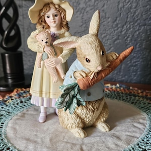 Könnte beinhalten: Eine Porzellanfigur eines Mädchens in einem gelben Kleid und einem Strohhut, das einen Teddybär hält, steht neben einem Kaninchen, das eine Karotte hält. Die Figur steht auf einer weißen Spitze mit einem blauen Häkelrand.