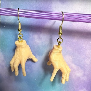 Puede incluir: Un par de pendientes de aro dorados con una escultura de mano de aspecto realista en un tono de piel claro. Las manos están en posición de 'paz'.