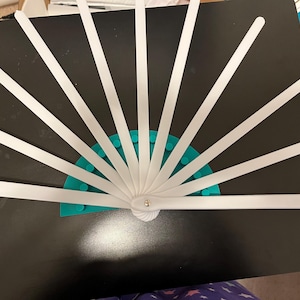 Może przedstawiać: Biały przedmiot w kształcie wachlarza z wieloma płaskimi, białymi paskami promieniującymi z centralnego punktu. Podstawa to półkole z turkusowego plastiku z małymi, okrągłymi wypustkami. Wachlarz jest ustawiony na ciemnej powierzchni.