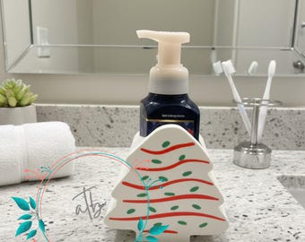 May include: A white ceramic Christmas tree-shaped soap dispenser holder with red and green accents. A dark blue soap bottle with a white pump sits inside the holder. The holder is on a white countertop.