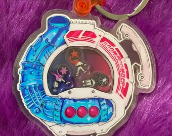 May include: A clear acrylic keychain featuring a circular design with a blue mechanical pipe element on the left and a red and white frame on the right. Inside the center window are three small character figures. The text BOOM BOOM GER is printed.