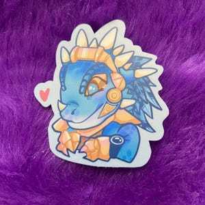 May include: A blue and gold cartoon dinosaur with a heart sticker. The dinosaur has a blue body, gold accents, and a white and blue feathered mane.