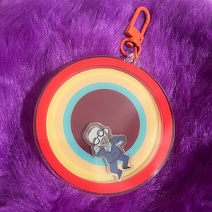 May include: A round keychain with a rainbow design and a cartoon figure. The keychain has a red border, with a clear center and an orange clasp. The cartoon figure is wearing a suit and glasses.