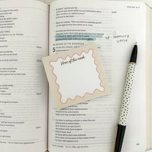 May include: A close-up of a pink sticky note with the text "Verse of the week" written in a white circle. The sticky note is placed on a page of an open Bible with the text "The Refuge of the Righteous" highlighted. The Bible is open to Psalms 4-5.