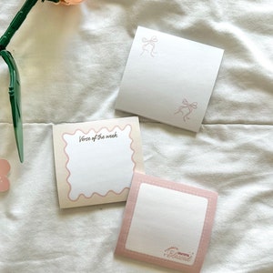 May include: Three pink and white notepads with different designs. One notepad has a scalloped edge and says "Verse of the week". Another notepad has a pink bow design. The third notepad has a pink border and says "Thankful Grateful Blessed".