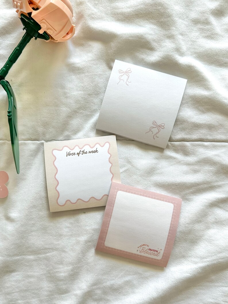 May include: Three pink and white notepads with different designs. One notepad has a scalloped edge and says "Verse of the week". Another notepad has a pink bow design. The third notepad has a pink border and says "Thankful Grateful Blessed".