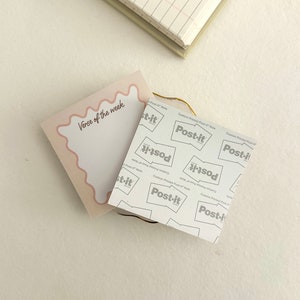 May include: A pink and white Post-it Note with the text "Verse of the week" written on it. The note is sitting on top of a stack of Post-it Notes with the brand name "Post-it" printed on them.