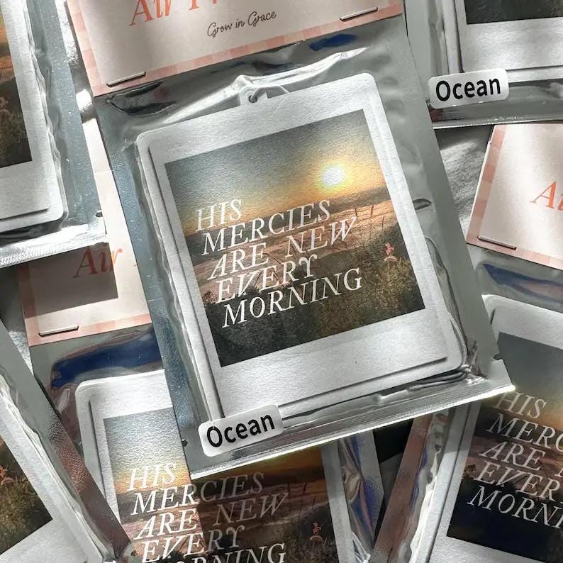 May include: A white car air freshener with a photo of a sunset and the text "His Mercies Are New Every Morning". The air freshener is labeled "Ocean".
