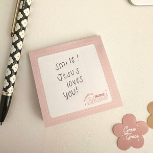 May include: A pink sticky note with the text "Smile! Jesus loves you!" and a small pink sticker with the text "Thankful, Grateful, Blessed" on a white surface. A black and white pen with a floral pattern is also visible.