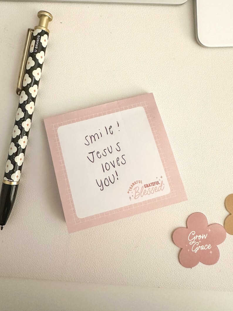 May include: A pink sticky note with the text "Smile! Jesus loves you!" and a small pink sticker with the text "Thankful, Grateful, Blessed" on a white surface. A black and white pen with a floral pattern is also visible.