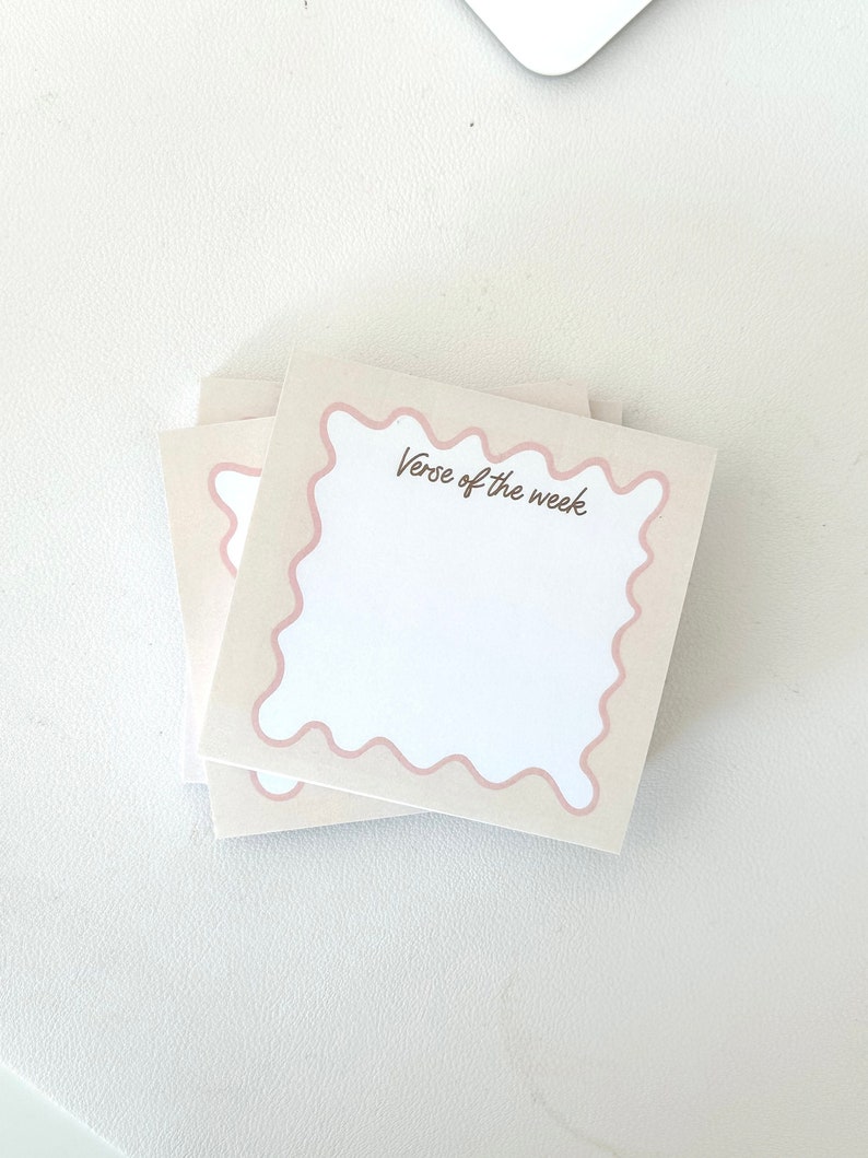 May include: A stack of pink and white notepads with a wavy border. The top notepad has the text "Verse of the week" written in brown.