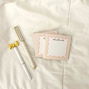 May include: Three pink and white notepads with rounded edges and a scalloped border. The top notepad has the text "Verse of the week" written in pink. Two pens, one white and one striped pink and white, are lying on a white surface.