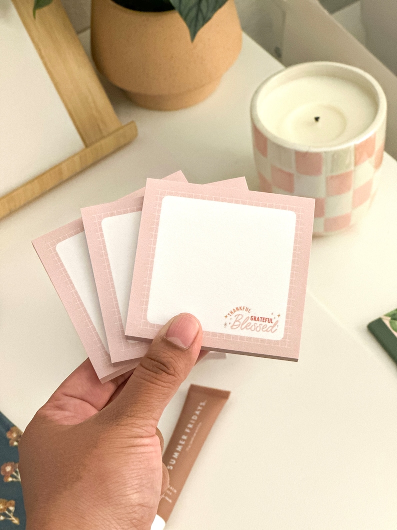 May include: Three pink sticky note pads with a grid pattern and a white writing surface. The top pad has the text "Thankful Grateful Blessed" in red and white.